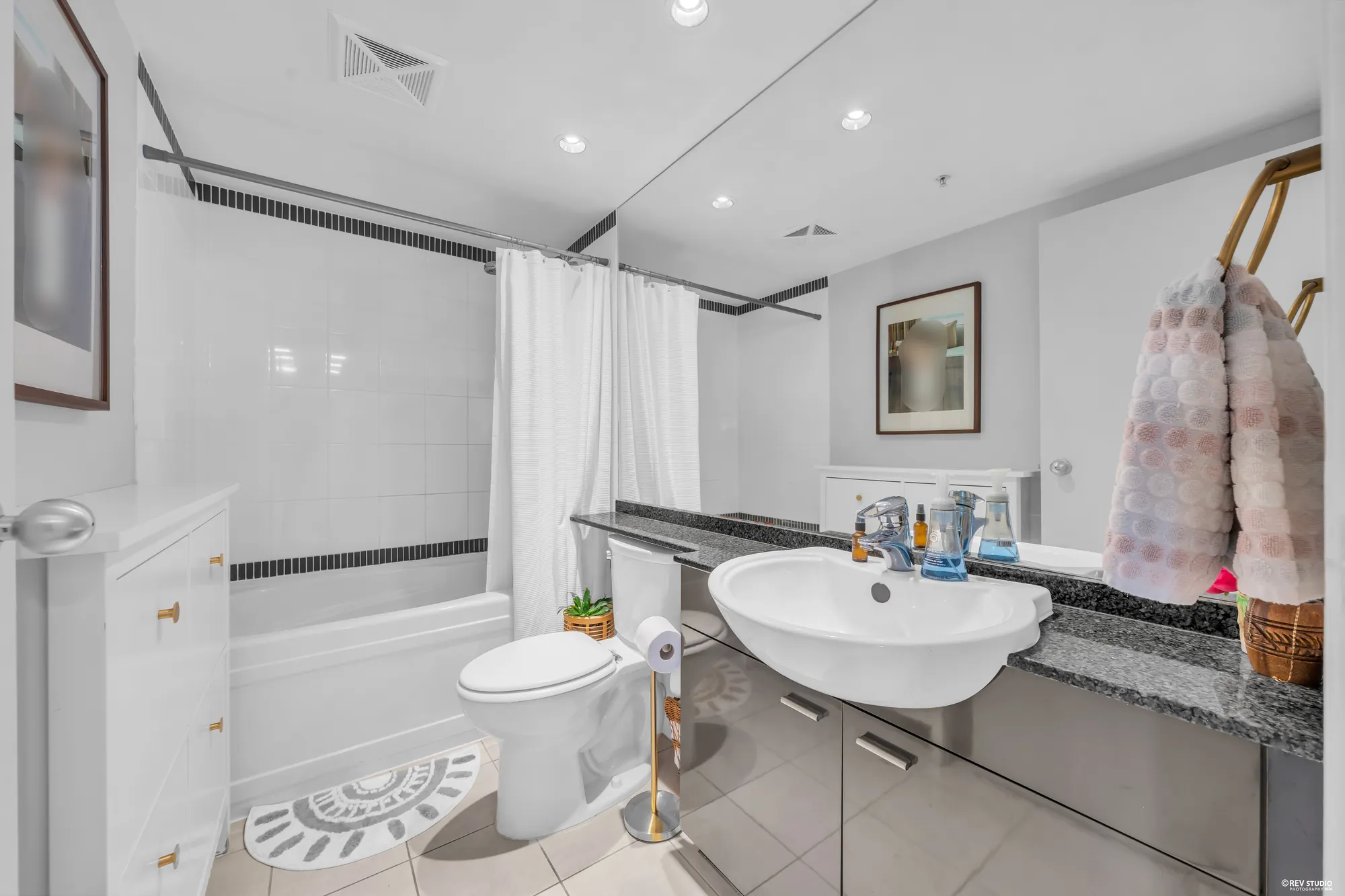 The bathroom: a tub with a white curtain, a basin on a black granite counter, a mirror wall, a framed photograph