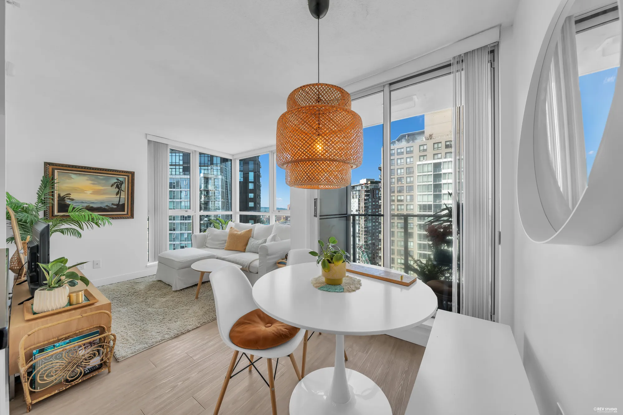 The dining table and chairs in the glass corner under a rattan pendant, a painting on the wall, the balcony and the view beyond
