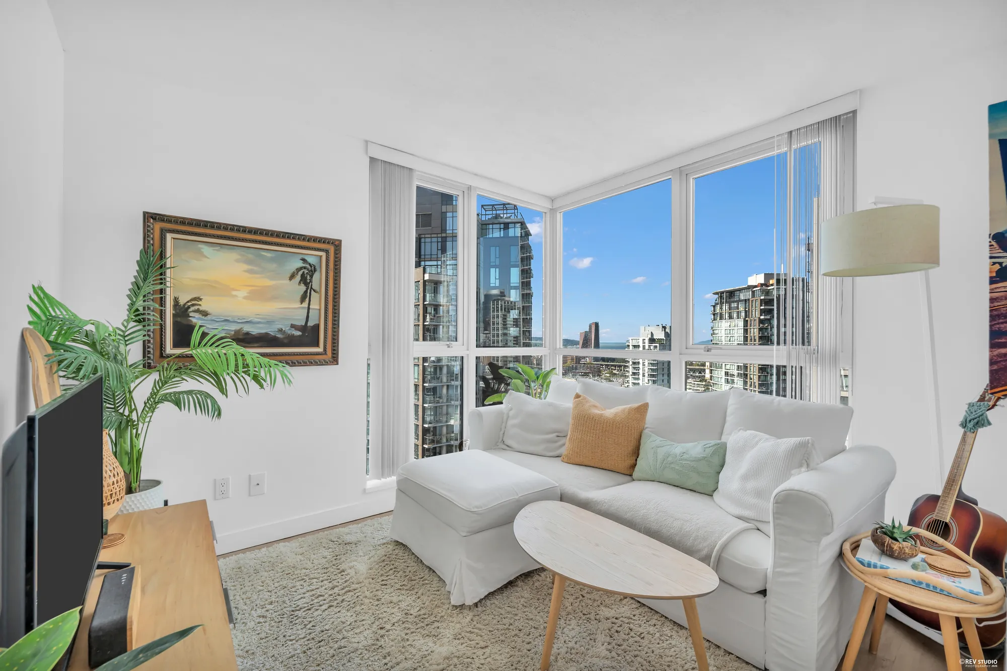 The living room: a white sofa and a low wooden console with a television, a palm, a painting on the wall, the corner of glass with the towers and the water beyond