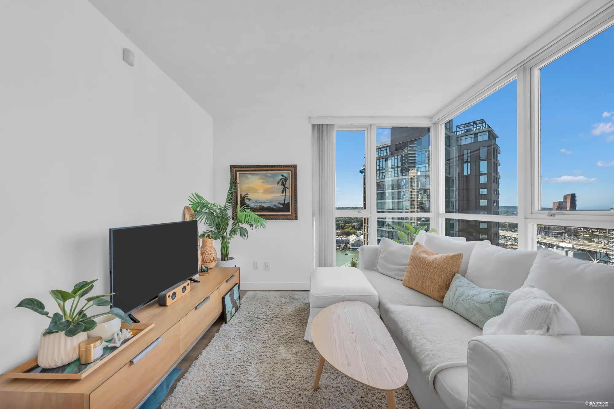 The living room of 3005-1408 Strathmore Mews: a white sofa and a television under a corner of floor-to-ceiling glass, the towers of Yaletown and False Creek beyond